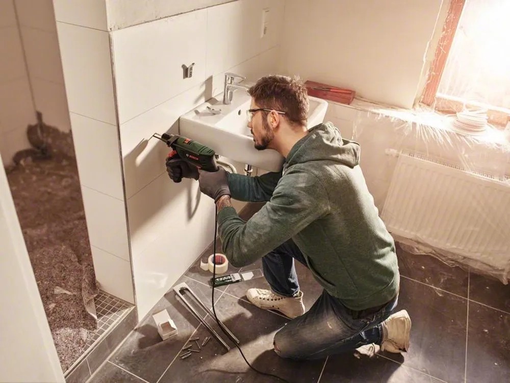 Muž vŕta do steny v kúpeľni s príklepovou vŕtačkou Bosch EasyImpact 5500. Muž vŕta do steny v kúpeľni s príklepovou vŕtačkou Bosch EasyImpact 5500.