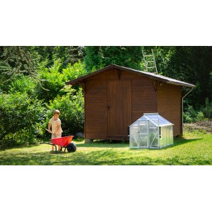 Mini skleník Vitavia Merlin vedľa záhradného domčeka, dieťa s fúrikom v záhrade.