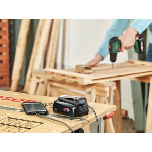 Bosch USB adaptér powerbanka nabíja smartfón na pracovnom stole; 18V akumulátorové náradie v dielni.