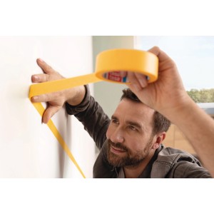 Muž lepí žltú tesa® Malerband Perfect+ na bielu stenu pre presné okraje maľby.