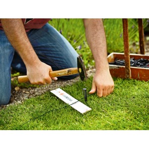 Inštalácia ohraničujúceho kábla Gardena: Muž upevňuje drôt pre robotickú kosačku kladivom v záhrade.