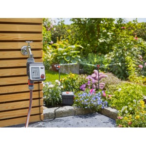 Zavlažovací počítač Gardena Master na kohútiku so zavlažovaním záhrady v pozadí.